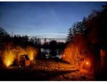 Abendliche Landschaft mit einem kleinen See im Hintergrund, umgeben von Bäumen und Büschen. Im Vordergrund sind brennende Beleuchtungen, die Büsche und eine kleine Steinstruktur orange anstrahlen, zu sehen.