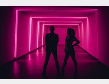 Ein moderner Gang mit beleuchteten Wänden und Decken in leuchtendem Pink, gestaltet durch geometrische Lichtstreifen. Im Vordergrund stehen die Silhouetten zweier Personen, die hinterleuchtet werden.