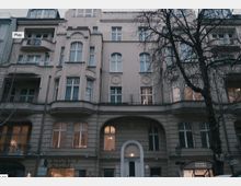 Fassade eines mehrstöckigen historischen Wohn- und Geschäftsgebäudes im urbanen Umfeld. Die Architektur zeigt verzierte Fensterrahmen, Rundbögen im Erdgeschoss und einen Eingang mit beleuchtetem Türbogen; im unteren Bereich befindet sich ein Schild mit der Aufschrift "Fasanen Passage".