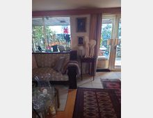 Wohnzimmer mit einer großen Glasfront, die auf einen Garten mit Pflanzen und Dekorationen blickt. Im Innenraum steht ein gemustertes Sofa, ein Beistelltisch mit Skulptur und Lampe, Teppiche auf Fliesenboden sowie dekorative Elemente wie Bilder und Kerzen.