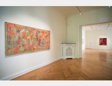 Helle Galerieräume mit hellem Holzparkett und weißen Wänden, an denen abstrakte, farbenfrohe Gemälde hängen. Im Vordergrund ein großes Kunstwerk, im Hintergrund ein weiterer Raum mit weiterer Kunst ausgeleuchtet.