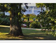 Ein gepflegter Park mit Blumenbeeten, Grünflächen und einem zentralen Springbrunnen im Vordergrund. Im Hintergrund ist ein Fluss mit einer Brücke und einer hohen Wasserfontäne zu sehen, umgeben von bewaldeten Hügeln.