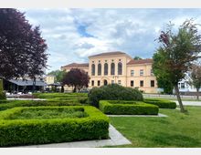 Gelbliches, historisches Gebäude mit großen Rundbogenfenstern und einem roten Ziegeldach, das von gepflegten Grünflächen mit Hecken, Büschen und Bäumen umgeben ist. Im Vordergrund befinden sich akkurat geschnittene Buchsbaumhecken, flankiert von einzelnen Laubbäumen.