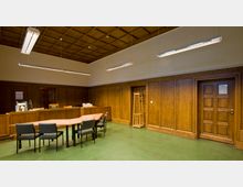 Holzvertäfeltes Büro oder Besprechungsraum mit einem grünen Boden, einer modernen Deckenbeleuchtung und einer Mischung aus klassischen Holzmöbeln, einschließlich eines geschwungenen Tisches und mehrerer Stühle. Zwei Türen und eine Garderobe sind in die mit Holz verkleideten Wände integriert.