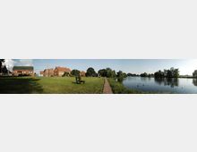 Panorama einer ländlichen Szenerie mit Grünflächen, einer Holzbank und einem schmalen Holzweg entlang eines Flusses. Im Hintergrund befinden sich Backsteingebäude und Bäume, die die Umgebung prägen.