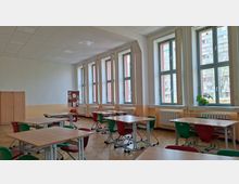 Heller Klassenraum mit mehreren Gruppentischen und rot-grünen Stühlen auf Parkettboden. An einer Wand befinden sich große Fenster mit Heizkörpern darunter, dazu ein Schrank und eine Pinnwand.