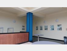 Innenansicht eines modernen Empfangs- oder Wartebereichs mit cremefarbenen Wänden, angewinkelten Deckenverkleidungen und blauer Säule. An der Wand hängen mehrere gerahmte Bilder, während im Vordergrund ein Tresen mit Holzoptik und runde Tische sichtbar sind.