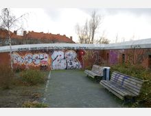 Eine kleine Parkanlage mit zwei Holzbänken und einem Mülleimer entlang eines gepflasterten Weges. Im Hintergrund steht eine mit Graffiti bedeckte Backsteinmauer, hinter der teilweise Dächer von Wohngebäuden und kahle Bäume sichtbar sind.