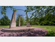 Eine kreisförmige Skulptur mit drei großen, rauen Steinsäulen, die ein Drahtgitterdach tragen, umgeben von grünen Bäumen und einem Weg aus rotem Kies in einem Park. Im Hintergrund sind Pflanzenbeete und weitere Grünflächen erkennbar.