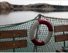 Ein ruhiger Gewässerabschnitt mit sandigen Hügeln im Hintergrund, gesehen von einem Boot mit Holzsitzen und einem grünen Netzgeländer, an dem ein rot-weißer Rettungsring befestigt ist.