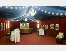 Innenansicht eines eleganten Veranstaltungsraums mit weinroten Wänden und einem blauen Stoffdach mit Lichterketten. Der Raum enthält zwei Stehtische mit weißen Tischdecken, eine geschwungene Bar mit Holzverkleidung, Kronleuchter an der Decke und gerahmte Fotografien an den Wänden.