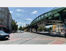 Straßenkreuzung in einer Stadt mit sichtbaren Straßenbahnschienen, Autos und Fahrrädern; ein grünes Hochbahn-Viadukt verläuft über die Straße. Im Hintergrund sind Bäume, Gebäude und der Berliner Fernsehturm sichtbar.