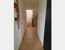 Ein moderner Flur mit hellem Holzboden, weißen Wänden und minimalistischer Gestaltung. Am Ende des Flurs führt eine gerade Treppe mit Holzstufen und einem Geländer aus Glas und Metall nach oben.