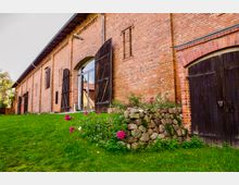 Backsteingebäude mit großen, dunkelbraunen Holztoren und einem Grünbereich im Vordergrund, der mit Blumenbeeten und einer kleinen Steinmauer gestaltet ist.