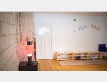 Eingangsbereich eines Gemeinschaftsraums mit Holzdielenboden, weißer Tür und einem niedrigen Schuhregal entlang der Wand. An der Wand steht „KIEZ-SPACE“ in bunten Buchstaben; links sind freigelegte, raue Mauerflächen und eine leuchtende Lampe zu sehen.