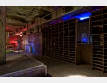 Industrieähnlicher Raum mit roher Betonoptik, großen Lüftungsschächten an der Decke und einer Wand mit vielen offenen Regalfächern. Der Raum ist spärlich beleuchtet mit farbigen Lichtern in Blau und Rot, was eine düstere Atmosphäre erzeugt.