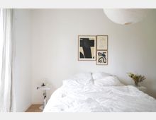 Minimalistisches Schlafzimmer mit einem weißen Bett, drei abstrakten Kunstwerken an der Wand und einem kleinen Nachttisch mit Büchern und Dekoration. Ein großer Papierlampenschirm hängt von der Decke, und eine Vase mit Trockenblumen steht auf einem Schrank rechts neben dem Bett.