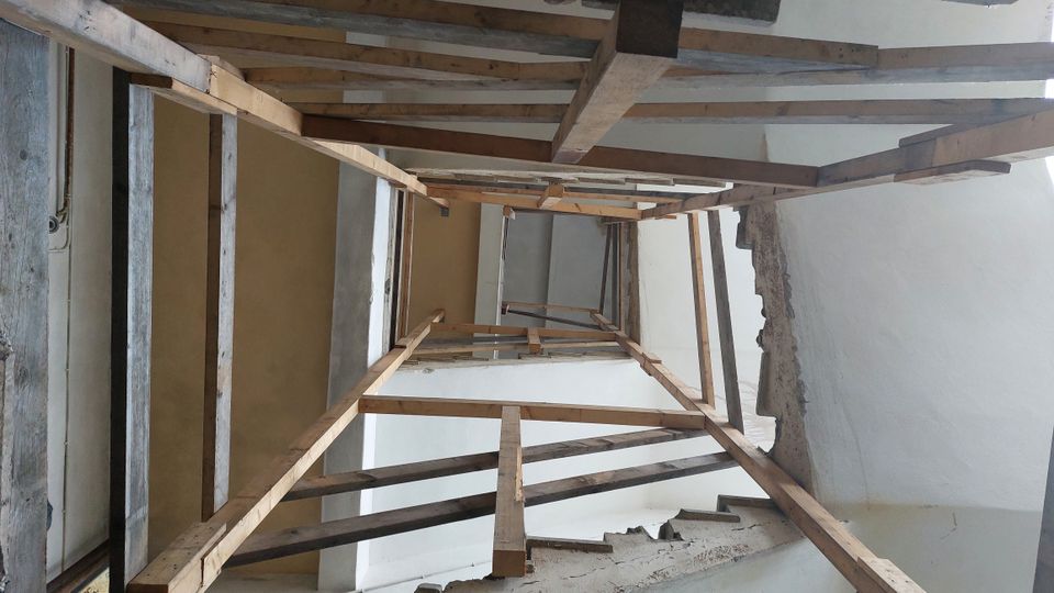 Holzgerüst in einem Gebäudetreppenhaus, mit freiliegenden Wänden aus weißem Putz und teilweise beschädigtem Mauerwerk. Das Gerüst wird verwendet, um Zugang bei Bau- oder Renovierungsarbeiten zu ermöglichen.
