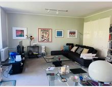 Modernes Wohnzimmer mit hellgrauen Wänden, großem schwarzen Ecksofa mit vielen Kissen und Glas-Couchtisch auf grauem Teppich. An der Wand hängen mehrere gerahmte Kunstbilder; links steht ein TV-Möbel vor einem Fenster, rechts sind hohe weiße Einbauschränke und ein Regal zu sehen.