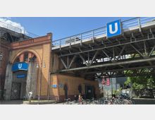 Eingangsbereich des U-Bahnhofs Gleisdreieck in Berlin mit einer Fassade aus gelbem Klinker und markanten roten Bögen, überdacht von einer erhöhten U-Bahn-Stahlkonstruktion. Im Vordergrund sind abgestellte Fahrräder und mehrere Hinweisschilder sichtbar.