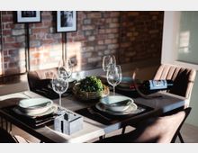 Ein modern gedeckter Esstisch mit vier Plätzen, ausgestattet mit hellen Tellern, Gläsern, Besteck und einer dekorativen Pflanze in einem flachen Korb in der Mitte. Im Hintergrund ist eine Wand aus rotem Backstein sowie zwei gerahmte Schwarz-Weiß-Bilder zu sehen.