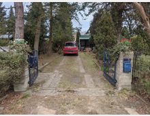 Blick durch ein geöffnetes blaues Metalltor zwischen zwei Betonpfeilern auf eine schmale Einfahrt mit Betonspuren, gesäumt von Bäumen und Sträuchern. Am Ende der Einfahrt steht ein Carport mit grünem Dach, davor ist ein rotes Auto geparkt.