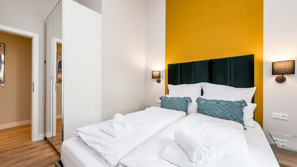 Modernes Schlafzimmer mit Doppelbett, weißer Bettwäsche und gerollten Handtüchern vor einer senfgelben Akzentwand mit gepolstertem dunklem Kopfteil. Seitlich stehen ein hoher weißer Kleiderschrank mit Spiegeltür, zwei Wandlampen und ein kleiner Glastisch auf Holzboden.
