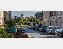 Kopfsteinpflasterstraße in einem Vorort mit parallel geparkten Autos und zweigeschossigen Wohnhäusern auf der rechten Seite. Im Hintergrund ist eine große Turmuhr von einem Industriebau sowie Grünflächen und Bäume zu sehen.