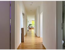 Ein heller Flur in einem modernen Gebäude mit Holzboden, weißen Wänden und mehreren Türen zu angrenzenden Räumen. Am Ende des Flurs steht eine Glastür, die zu einem Bereich mit Pflanzen und Tageslicht führt.