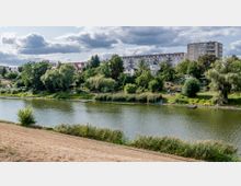 Ufer einer ruhigen Flusslandschaft mit dichter Vegetation und kleinen Booten, im Hintergrund eine Reihe von Wohnhäusern, darunter ein kleines Mehrfamilienhaus und ein höheres Wohngebäude. Der Himmel ist teilweise bewölkt.