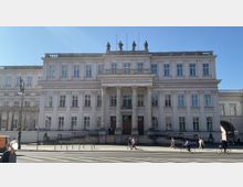 Neoklassizistisches Gebäude mit symmetrischer Fassade, einer Säulenfront am Eingangsbereich und mehreren Statuen auf dem Dach; im Vordergrund eine Straße mit Fußgängern und Radfahrern bei Sonnenschein.