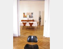 Ein heller Innenraum mit Holzparkettboden, einem Esstisch mit weißer Tischdecke und mehreren Holzstühlen. Im Hintergrund stehen ein Sideboard, darauf dekorative Gegenstände, und an der Wand hängen zwei bunte geometrische Bilder. Im Vordergrund ist ein schwarzer Stuhl sichtbar.