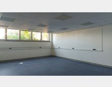 Leerstehender Raum mit weiß gestrichenen Wänden, heller Rasterdecke mit eingebauten Leuchtkästen und großen Fenstern, die Tageslicht hereinlassen. Der Boden ist dunkelgrau, und entlang der Wände sind Kabelkanäle und Steckdosen angebracht.