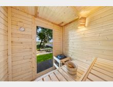 Innenraum einer kleinen Holzsauna mit hellen Holzvertäfelungen, Saunabänken, Saunaofen mit Steinen, Eimer und Thermometer. Durch die Glastür blickt man auf eine Grünfläche mit Baum und Sitzgelegenheit nahe einem Gewässer.