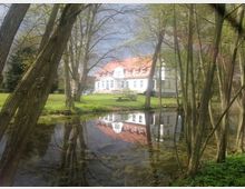Herrenhaus mit weißer Fassade und rotem Ziegeldach, umgeben von Rasen und Bäumen, reflektiert in einem kleinen ruhigen Teich im Vordergrund. Der Himmel ist bewölkt, die Umgebung wirkt naturnah.