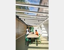 Ein moderner, heller Innenraum mit einem Glasdach, durch das Sonnenlicht einfällt. Im Raum steht ein langer Holztisch mit bunten Stühlen, an dem drei Personen arbeiten; die Wände sind teils aus Beton, teils mit grünen Wandpaneelen dekoriert.