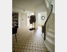 Heller Hausflur mit gemustertem Fliesenboden, weiß gestrichenen Wänden und einer Treppe mit dunklem Handlauf rechts. Links stehen ein offenes Schuhregal und eine Tür, in der Mitte eine Garderobe, hinten ein Fenster und ein Wandspiegel.