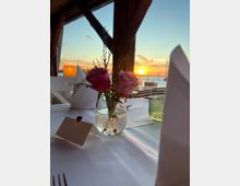 Ein festlich gedeckter Tisch in einem Restaurant mit Blick auf einen Sonnenuntergang über einem Gewässer. Im Vordergrund stehen ein Glas mit rosa Blumen, eine Tischkarte und eine grüne Glasvase, während durch große Holzfenster die Außenlandschaft mit Holzstegen und Spiegelungen der Sonne sichtbar ist.