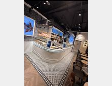 Moderner Eisladen mit geschwungener, weißer Verkaufstheke, darauf Speisen und digitale Displays. Der Raum hat dekorative Wandmuster, eine dunkle Decke mit Hängelampen, gemusterte Fliesenböden und eine Sitzbereich mit runden Tischen und Stühlen.