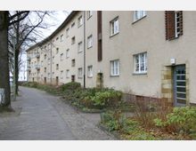 Mehrstöckiges Wohngebäude mit heller Fassade, rechteckigen Fenstern und vereinzelt sichtbaren Eingangstüren; davor ein Fußweg, gesäumt von Bäumen und niedrigen Sträuchern in einer gepflegten Grünanlage.