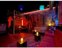 Ein gemütlicher, dunkel beleuchteter Lounge-Bereich mit rotem Sofa, cremefarbenen Sitzmöbeln und kleinen Tischen mit elektrischen Kerzen. Im Hintergrund befindet sich eine Holzkonstruktion mit Sitznische, umgeben von Perlenschnüren, sowie eine Wand mit abstrakter Kunst und dekorativen Details.