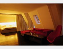 Modern eingerichtetes Interieur mit einem Schlafzimmerbereich, der durch Vorhänge abgetrennt ist, sowie einem Wohnbereich mit rotem Sofa, Sesseln und einem Couchtisch. Schrägwände und ein kleines Dachfenster prägen die Architektur, während eine warme Beleuchtung den Raum erhellt.