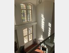 Innenansicht eines hohen Raums mit weißen Wänden und großen, mehrfach unterteilten Bogenfenstern, die Tageslicht einlassen. Am Boden befinden sich rotbraune Fliesen, an der Wand ein Heizkörper und ein Kamin, teilweise von Schatten beleuchtet.