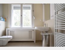 Helles Badezimmer mit cremefarbenen Fliesen, einem Fenster mit Blick ins Grüne, einem Waschbecken mit Spiegel, einer Toilette und einem weißen Handtuchheizkörper.