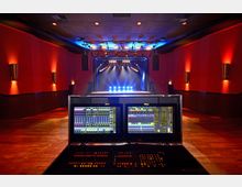Ein Konzertsaal mit roter Wandverkleidung und dimmbarer Beleuchtung. Im Vordergrund befindet sich ein Mischpult mit zwei leuchtenden Bildschirmen, dahinter eine Bühnenfläche mit blauer und weißer Scheinwerferbeleuchtung.