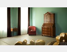 Ein Raum mit mintgrüner Wand, zwei großen Fenstern mit dunklen Vorhängen, einem roten Sessel, einer antiken Kommode aus Holz mit Schubladen und gewölbtem Abschluss sowie einem kleinen Beistelltisch. Der Boden ist betoniert, und im Vordergrund ist ein goldfarbener Stuhlteil sichtbar.