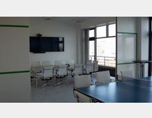 Moderner Konferenzraum mit weißen Bürostühlen um einen großen rechteckigen Tisch, einem großen Bildschirm an der Wand und Fensterfronten, die Tageslicht hereinlassen. Im Vordergrund steht ein Tischtennistisch, und an der Seite befindet sich ein kleiner Stehtisch.