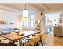 Helles Wohnzimmer mit offener Küche in einem modernen Apartment. Der Raum verfügt über einen Holztisch mit Stühlen, eine helle Sitzecke mit Sofa, große Fenster mit weißen Vorhängen und eine Backsteinwand mit Dekorationen; im Hintergrund ist eine kompakte Küche mit weißen Fliesen sichtbar.