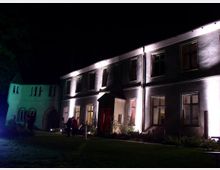 Nachtszene eines historischen Gebäudes mit zweistöckiger Fassade, beleuchtet durch Strahler, die helle Fenster und eine zentrale Eingangstür hervorheben. Im Vordergrund befindet sich eine Rasenfläche, und links im Hintergrund ist ein kleinerer, turmähnlicher Abschnitt des Gebäudes sichtbar.