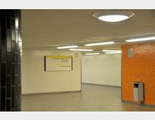 U-Bahn-Unterführung mit hellen Wänden, einer orangefarbenen Wand mit Mosaikfliesen und einem Wandmülleimer. Ein Wegweiser zeigt Richtungen zu Ausgängen und Anschlussmöglichkeiten an.